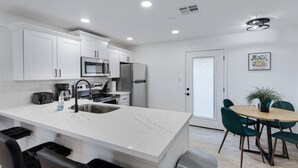 A modern kitchen with white cabinets, stainless steel appliances, a marble countertop, and a dining area with a wooden table and green chairs. - Weber · Jungle House, minutes away from ASU/ Old Town (Tempe)