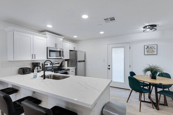 A modern kitchen with white cabinets, stainless steel appliances, a marble countertop, and a dining area with a wooden table and green chairs.