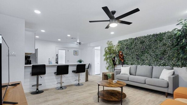 A modern living room with a grey sofa, a coffee table, and two black bar stools. A ceiling fan is visible, and there is a green wall with plants. - Weber · Jungle House, minutes away from ASU/ Old Town (Tempe)