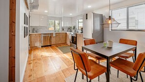 Une cuisine moderne avec un plancher en bois, un coin repas avec des chaises orange et une grande fenêtre équipée de stores.