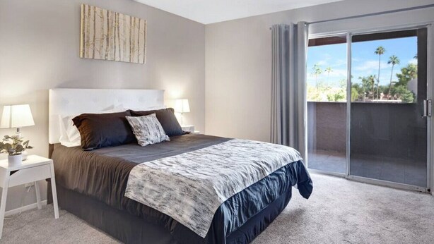 Un dormitorio moderno con una cama grande, una mesita de noche, una lámpara y vistas a palmeras a través de un ventanal corredero.