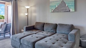 Un sofá seccional gris en una habitación con un cuadro de un muelle y el océano en la pared, una lámpara de pie y vistas a través de una puerta de cristal.