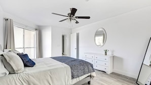 Et soveværelse med seng, ventilator, kommode og spejl.