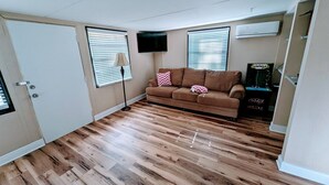 A compact living space with a brown sofa, wooden flooring, a flat-screen TV, and a ceiling-mounted air conditioner.