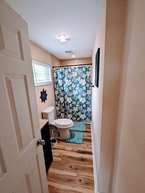 Een smalle badkamer met een toilet, een douchegordijn met een bloemenmotief en houten vloeren.