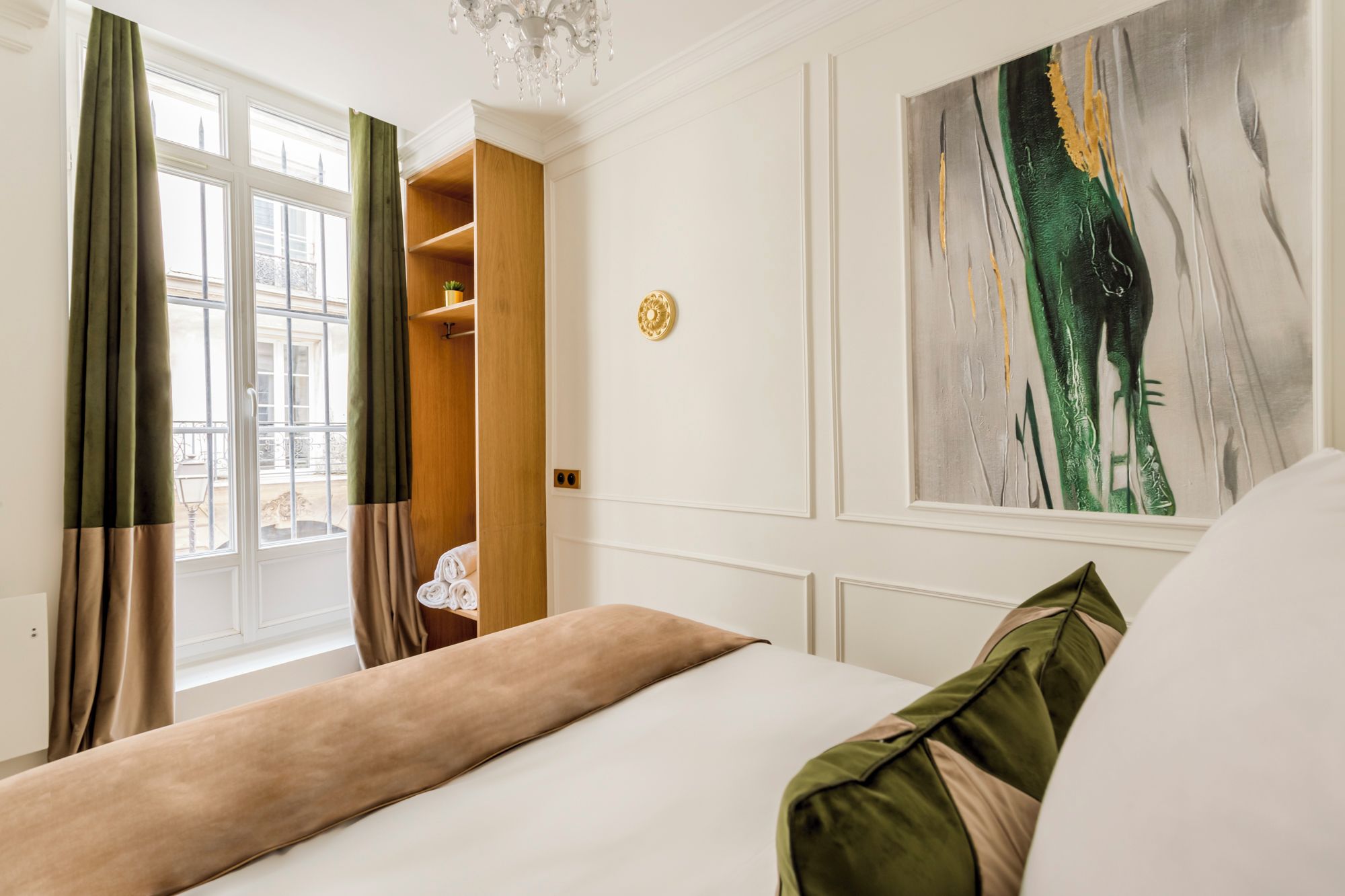 A hotel room with a bed, a wardrobe, a window with curtains, a chandelier, and a painting on the wall.