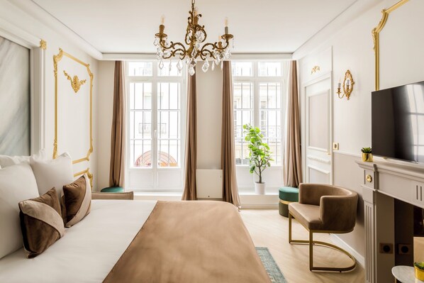 A spacious bedroom with a large bed, a sitting area, a television, and a view of the city through large windows.