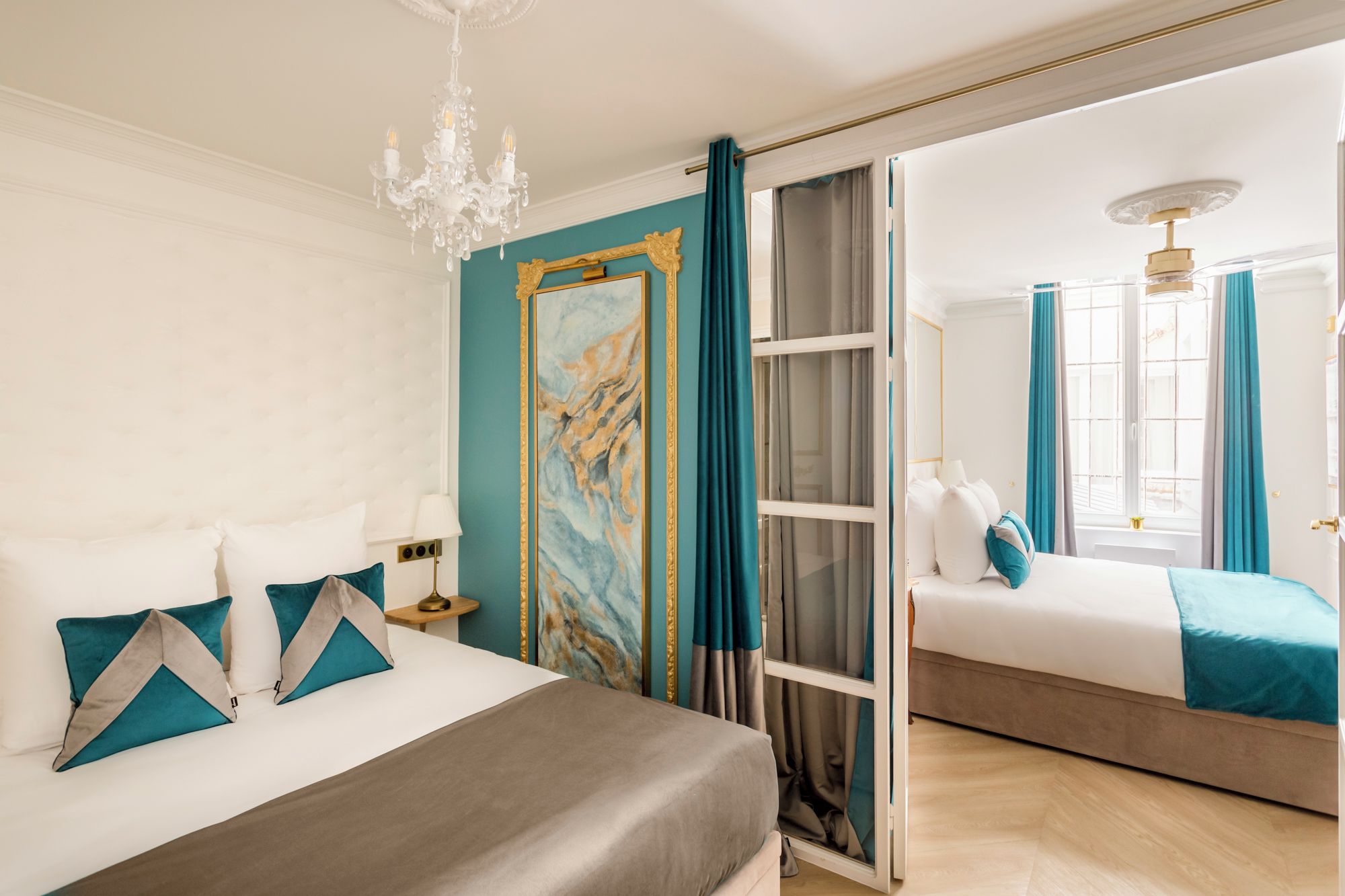 Une chambre d’hôtel avec un grand lit, un lit plus petit, un lustre, un tableau et une vue sur l’extérieur, visible à travers une porte vitrée.