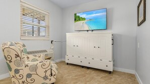 Una habitación con un armario blanco, un sillón floral y un televisor en la pared.
