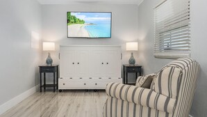 A bedroom with a bed, a TV mounted on the wall, a window with blinds, and a small table with a lamp. - 2-Bedroom Condo with screened patio - Manatee - Welcome to our newly renovated, spacious and luxurious two-bedroom condo! (Matlacha)