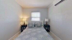 A bedroom with a bed, two bedside tables with lamps, a window with blinds, and an air conditioning unit. - 2-Bedroom Condo with screened patio - Manatee - Welcome to our newly renovated, spacious and luxurious two-bedroom condo! (Matlacha)