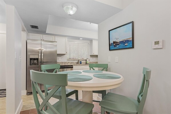 A modern kitchen with a stainless steel refrigerator, a dining table with four chairs, and a window with blinds.