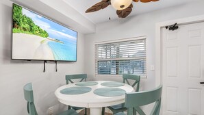 Un comedor con una mesa redonda, cuatro sillas, un ventilador de techo y un televisor mostrando una escena de playa.