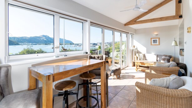 Um quarto espaçoso com uma grande janela, vigas de madeira no teto, uma mesa de madeira com banquinhos, móveis de vime e vista para um corpo de água e montanhas.