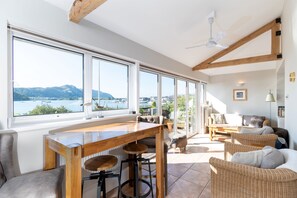 Een ruime kamer met een groot raam, houten balken in het plafond, een houten tafel met krukken, rieten meubels, en uitzicht op een waterlichaam en bergen.