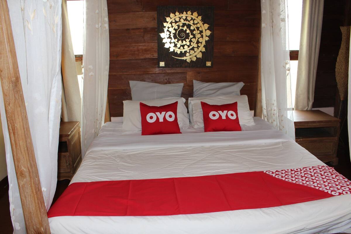 Ein Bett mit weißer Bettwäsche und zwei roten Kissen mit dem Text 'OYO', ein hölzernes Kopfteil und ein Wandbild darüber.