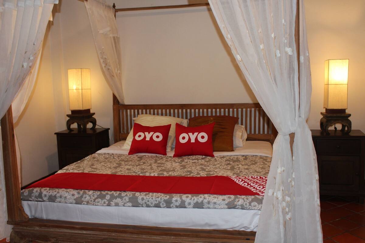 Ein Himmelsbett mit Baldachin, zwei Nachttischlampen und Kissen mit 'OYO'-Branding.