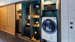 A compact laundry area with a front-loading washing machine, a small trash bin, and a shelf with various items. - Sumiya Seica (Takayama)