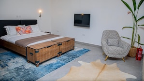 A modern bedroom with a bed, a wooden bench, a patterned armchair, a large plant, and a fire extinguisher. - CASA BIANCA (Accra)