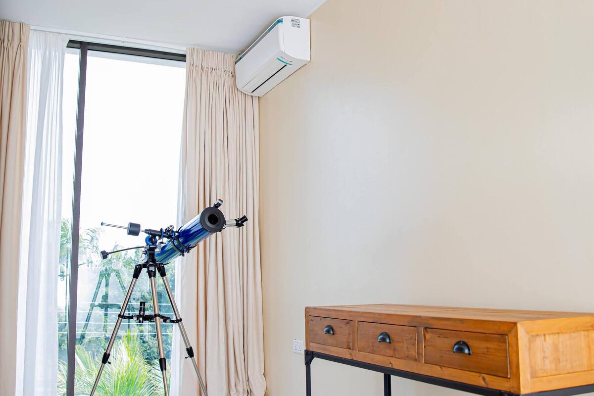 A room with a telescope on a tripod, a wooden table with drawers, a vase, and an air conditioning unit on the wall.