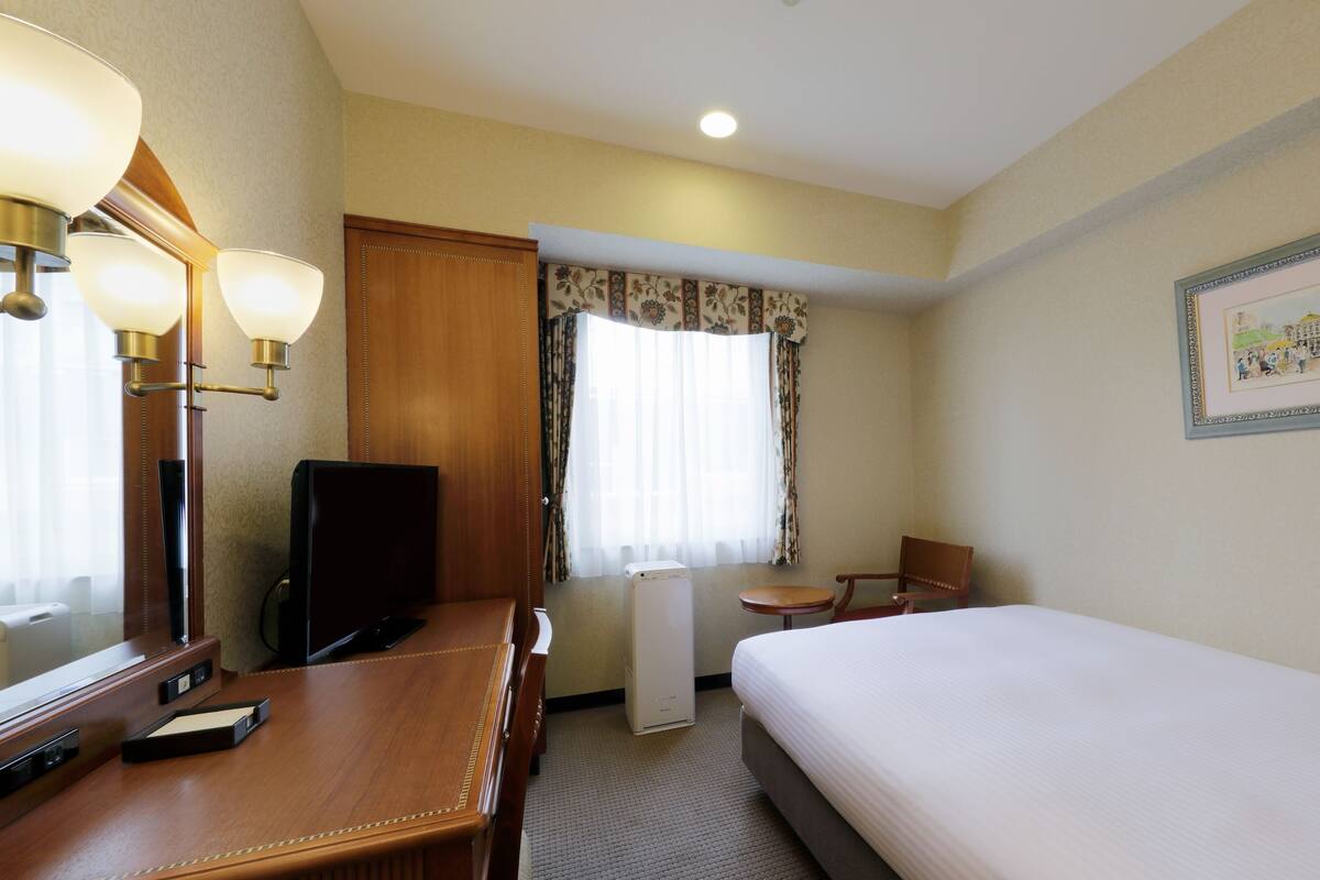 Habitación de hotel con cama, escritorio con computadora, silla, televisor, mesita y ventana con cortinas. En la pared, un cuadro enmarcado.