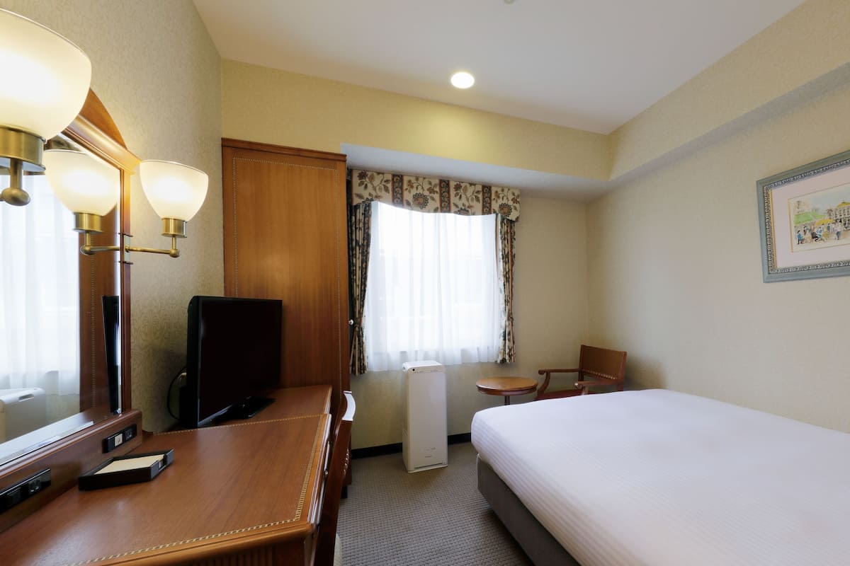 A hotel room with a bed, a desk with a computer, a chair, a television, a small table, a window with curtains, and a framed picture on the wall.