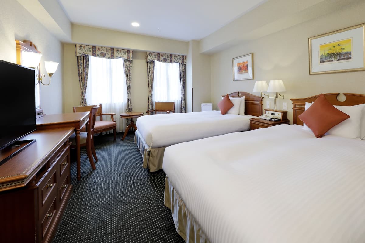 A hotel room with two beds, a desk with a television, a chair, a small table, and two lamps.