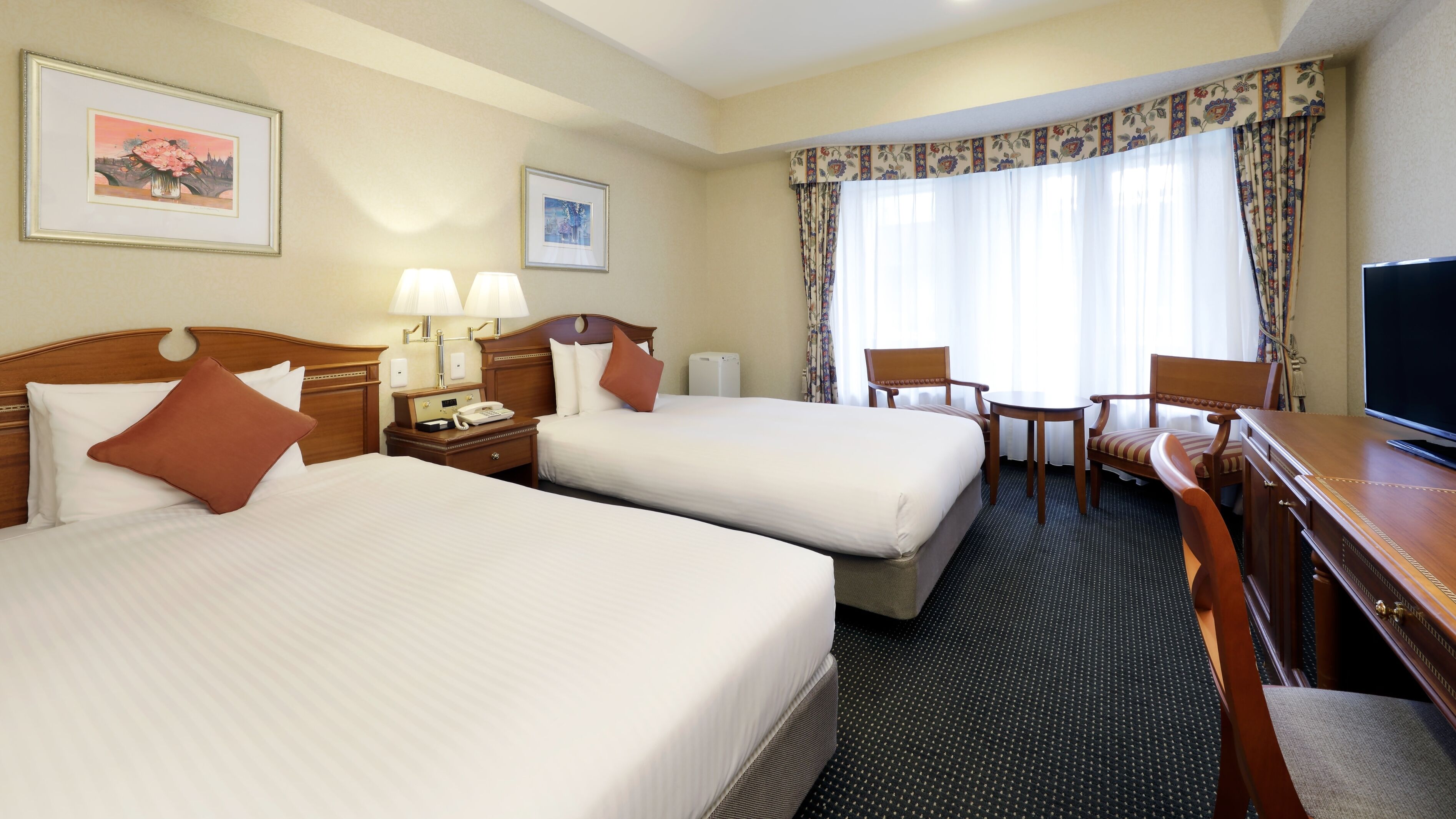 A hotel room with two beds, a desk, a chair, a television, and a window with curtains.