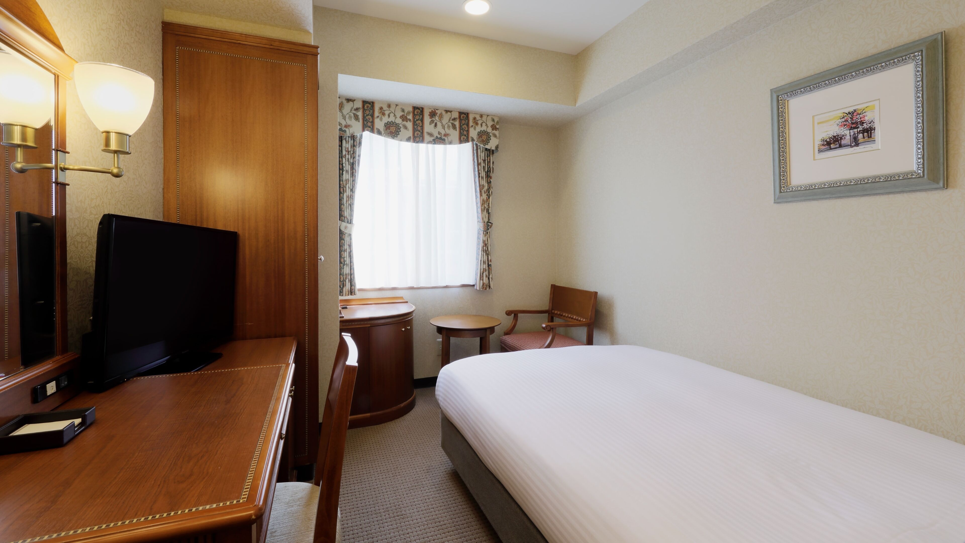 A hotel room with a bed, a desk with a television, a chair, a small table, and a window with curtains.