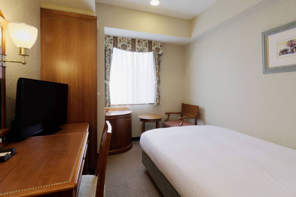 Habitación de hotel con cama, escritorio con televisor, silla, mesita y ventana con cortinas.
