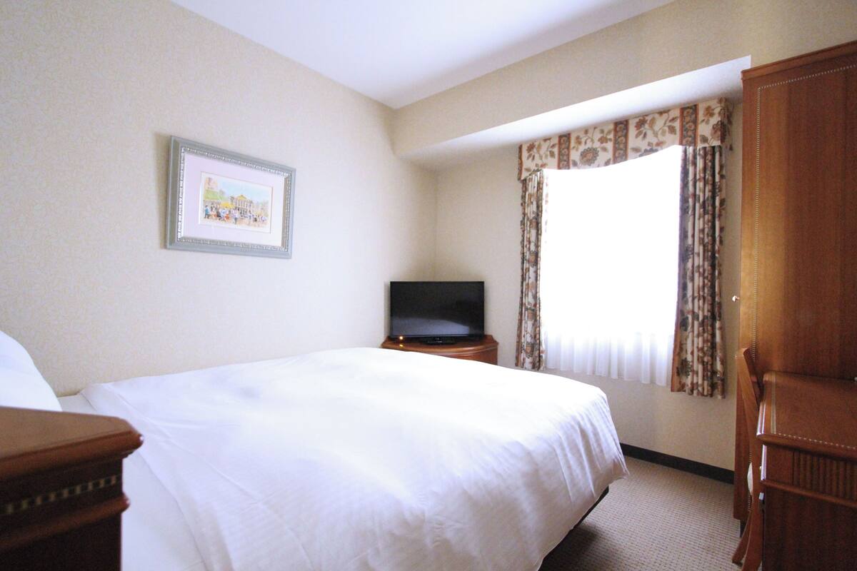 Habitación de hotel con una cama grande, un televisor sobre un soporte de madera, un cuadro en la pared y una ventana con cortinas.