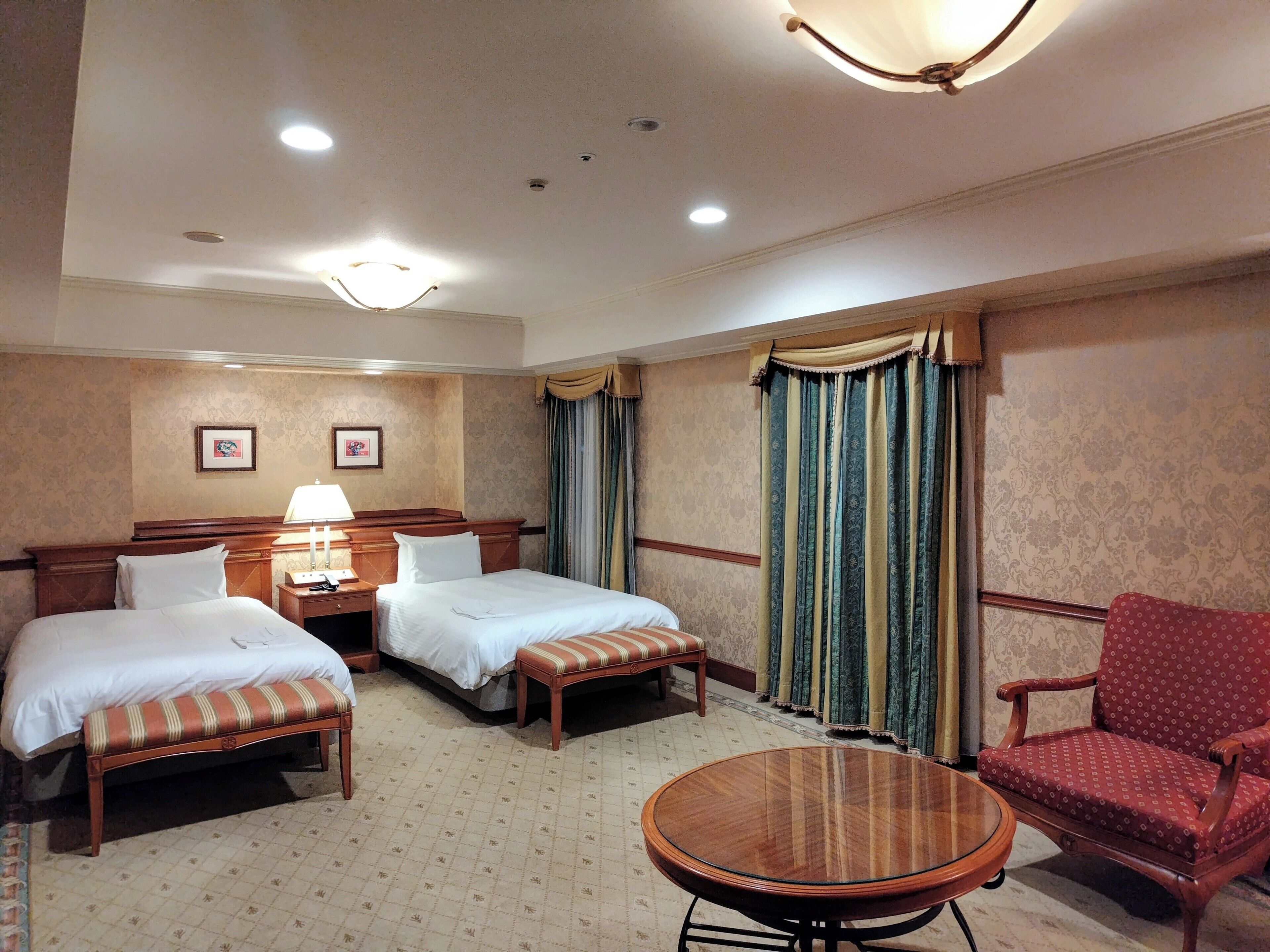 A hotel room with two beds, a chair, a round table, and a lamp.