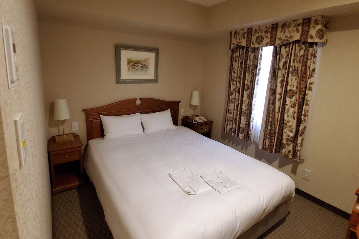Habitación de hotel con una cama grande, dos mesitas de noche, un buró, un cuadro enmarcado en la pared y una ventana con cortinas.