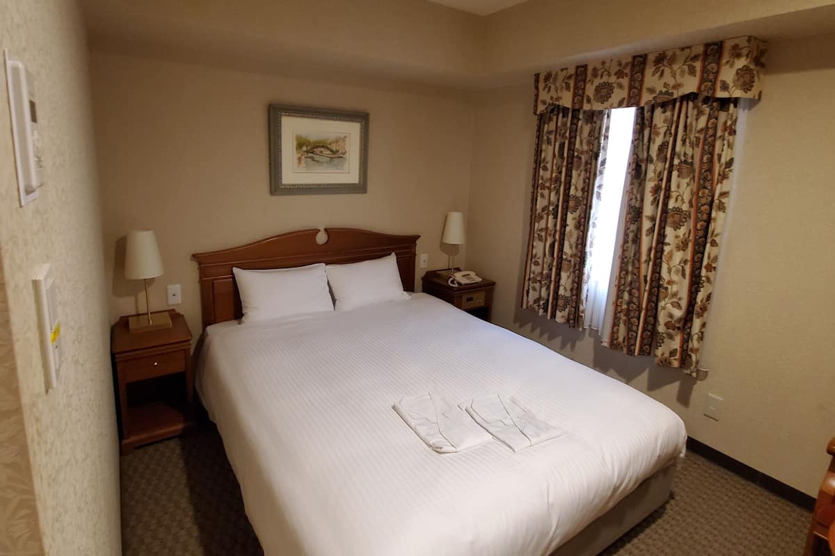 A hotel room with a large bed, two bedside tables, a nightstand, a framed picture on the wall, and a window with curtains.
