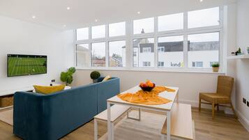 A modern living room with a large window, a flat-screen TV, a blue sofa, a wooden table with a bowl of fruit, and a wicker chair.