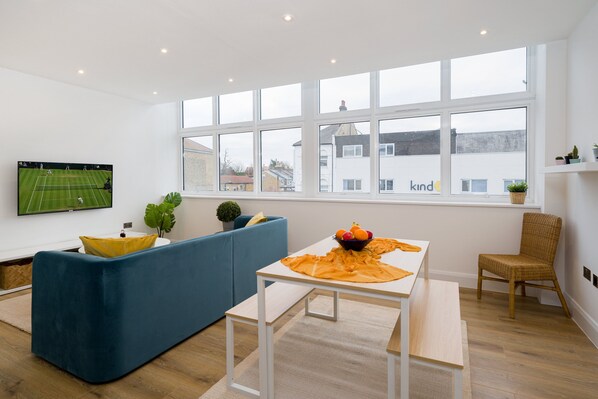 A modern living room with a large window, a flat-screen TV, a blue sofa, a wooden table with a bowl of fruit, and a wicker chair. - Panther House by Oval (London)