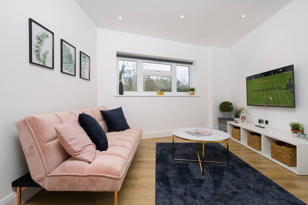 A modern living room with a pink sofa, a round coffee table, and a large TV screen displaying a tennis match.