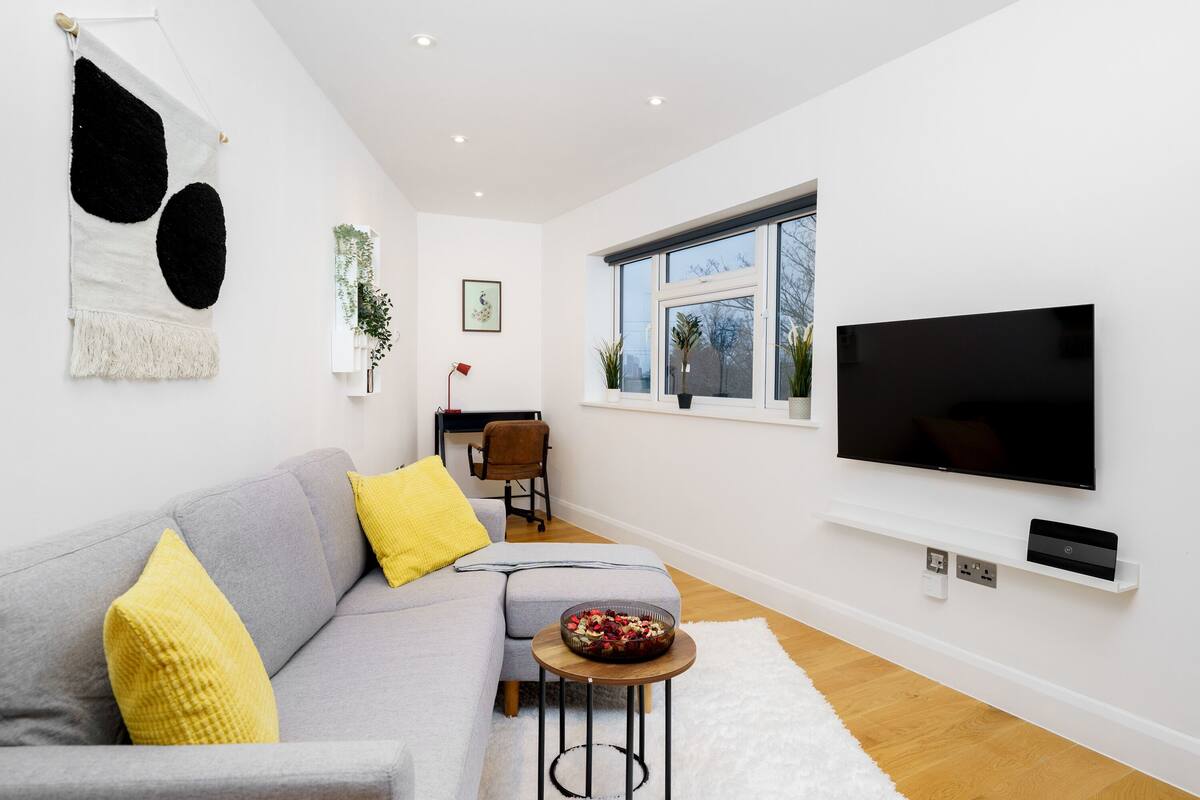 A modern living room with a grey sofa, yellow cushions, a round coffee table, and a large flat-screen TV mounted on the wall.