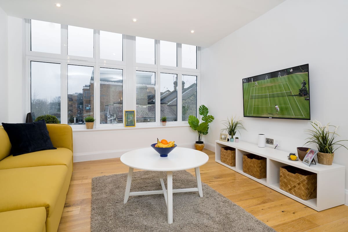 A modern living room with a yellow sofa, a white coffee table with fruit, a TV on the wall, and a large window with city views.