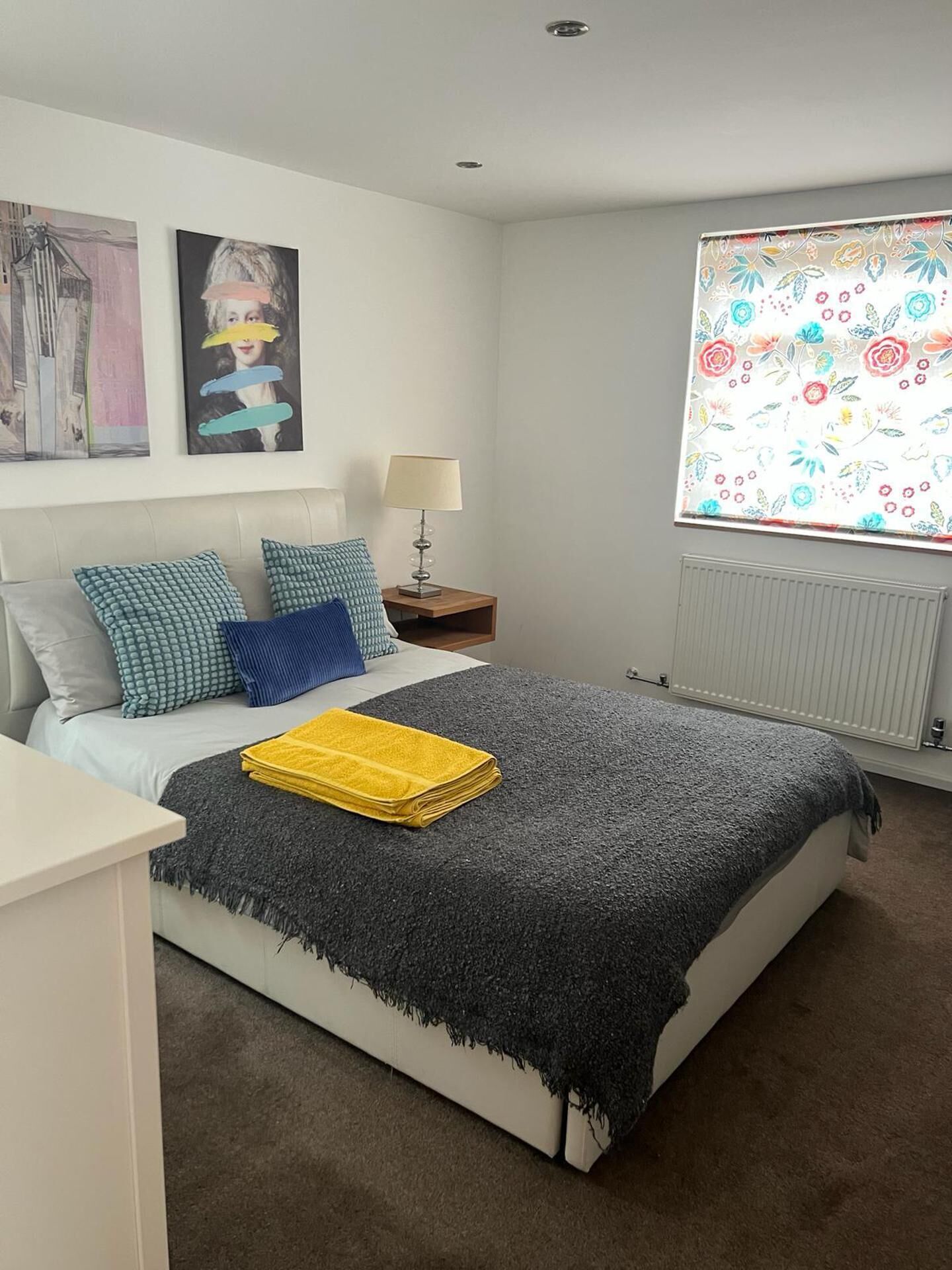 Une chambre à coucher avec un lit, des oreillers, une table de chevet avec une lampe, une fenêtre ornée d’un motif floral et des œuvres d’art accrochées au mur.
