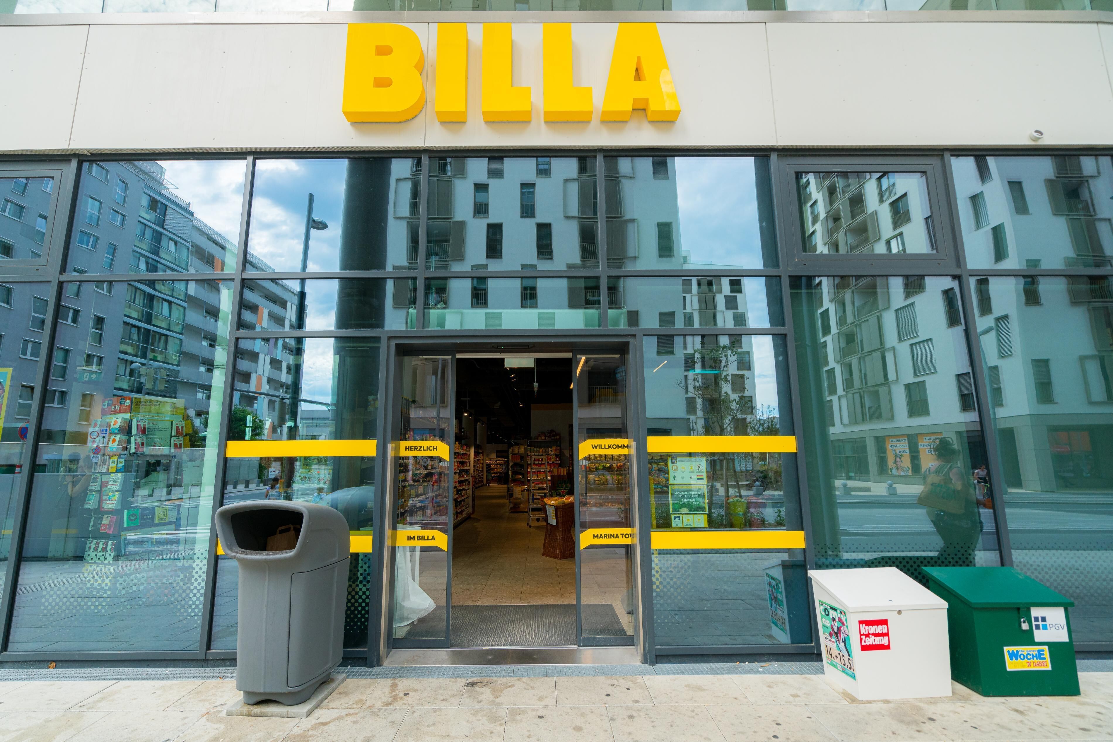 L’entrée d’un magasin BILLA, avec des portes vitrées, une poubelle et une poubelle de recyclage verte à l’extérieur.