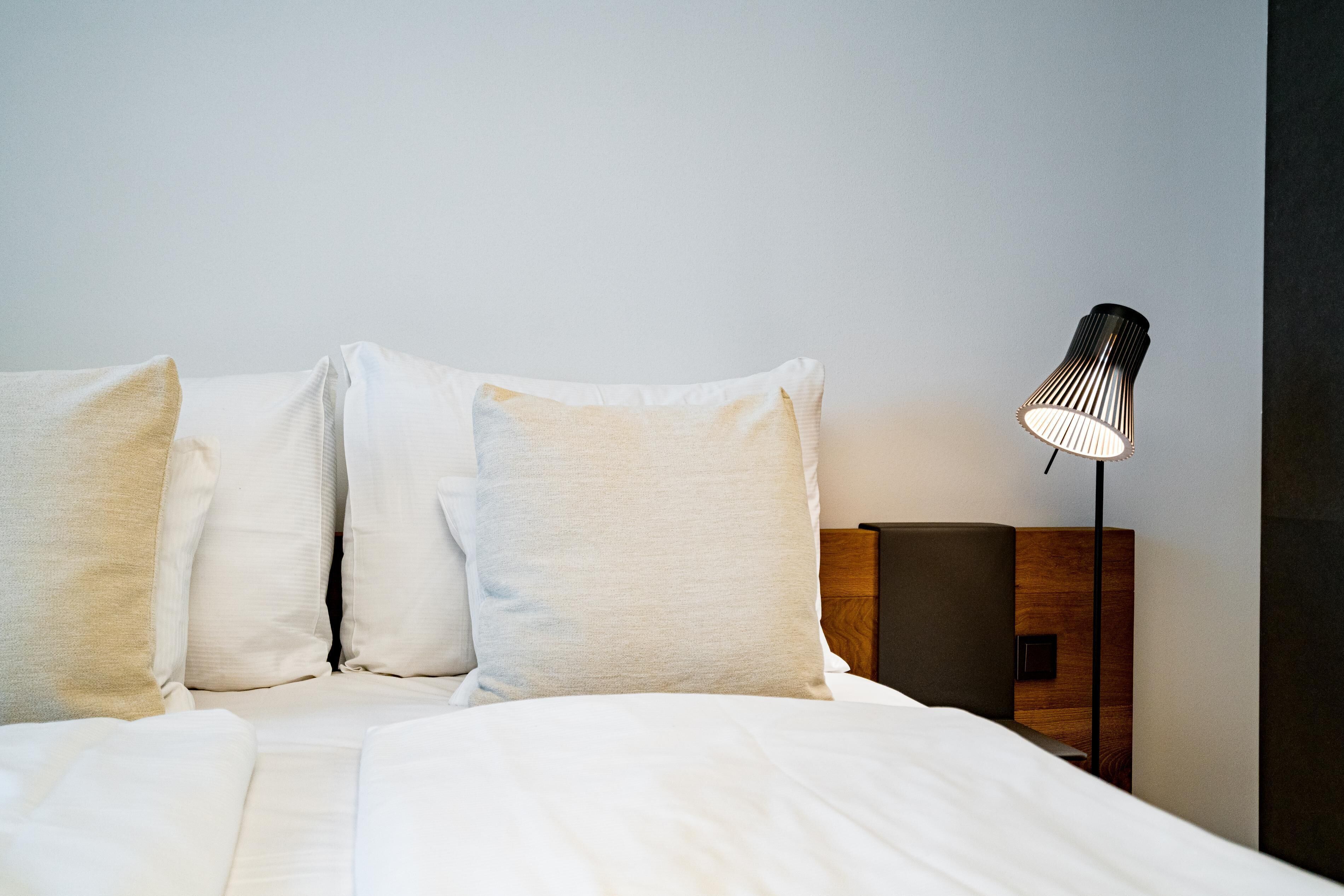 A neatly made bed with white linens and three pillows, a modern lamp on a wooden nightstand, and a plain wall in the background.