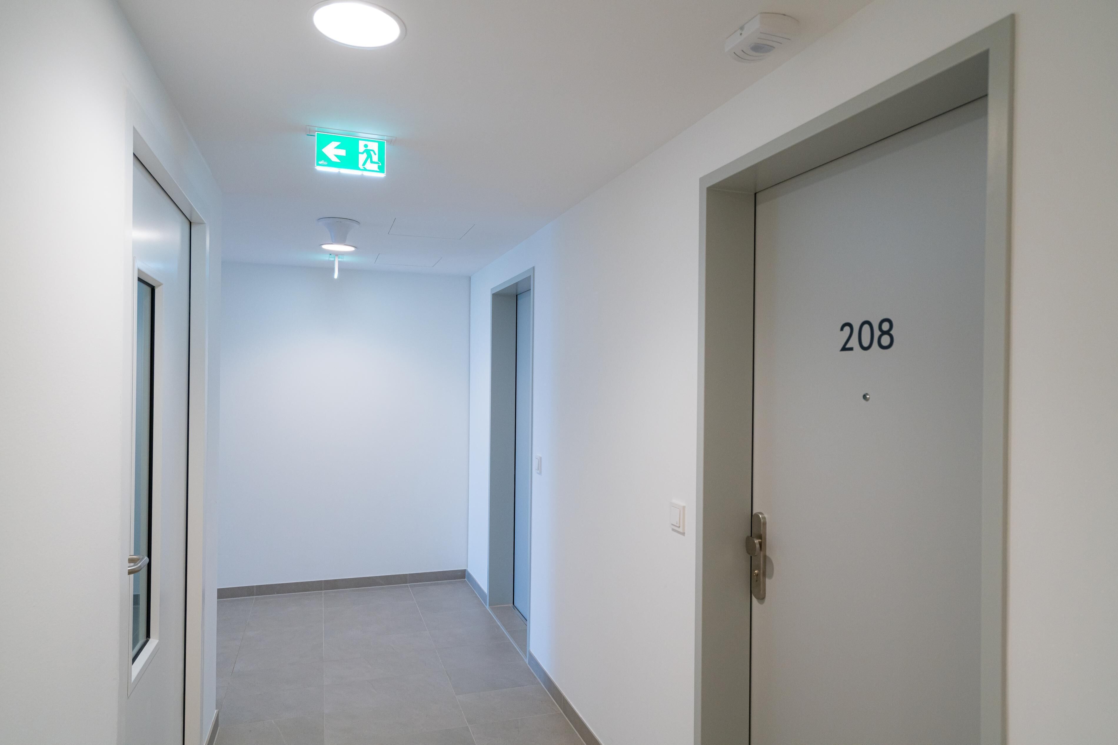 Un pasillo con una puerta marcada con el número 208, un interruptor de luz y un letrero verde de salida de emergencia.