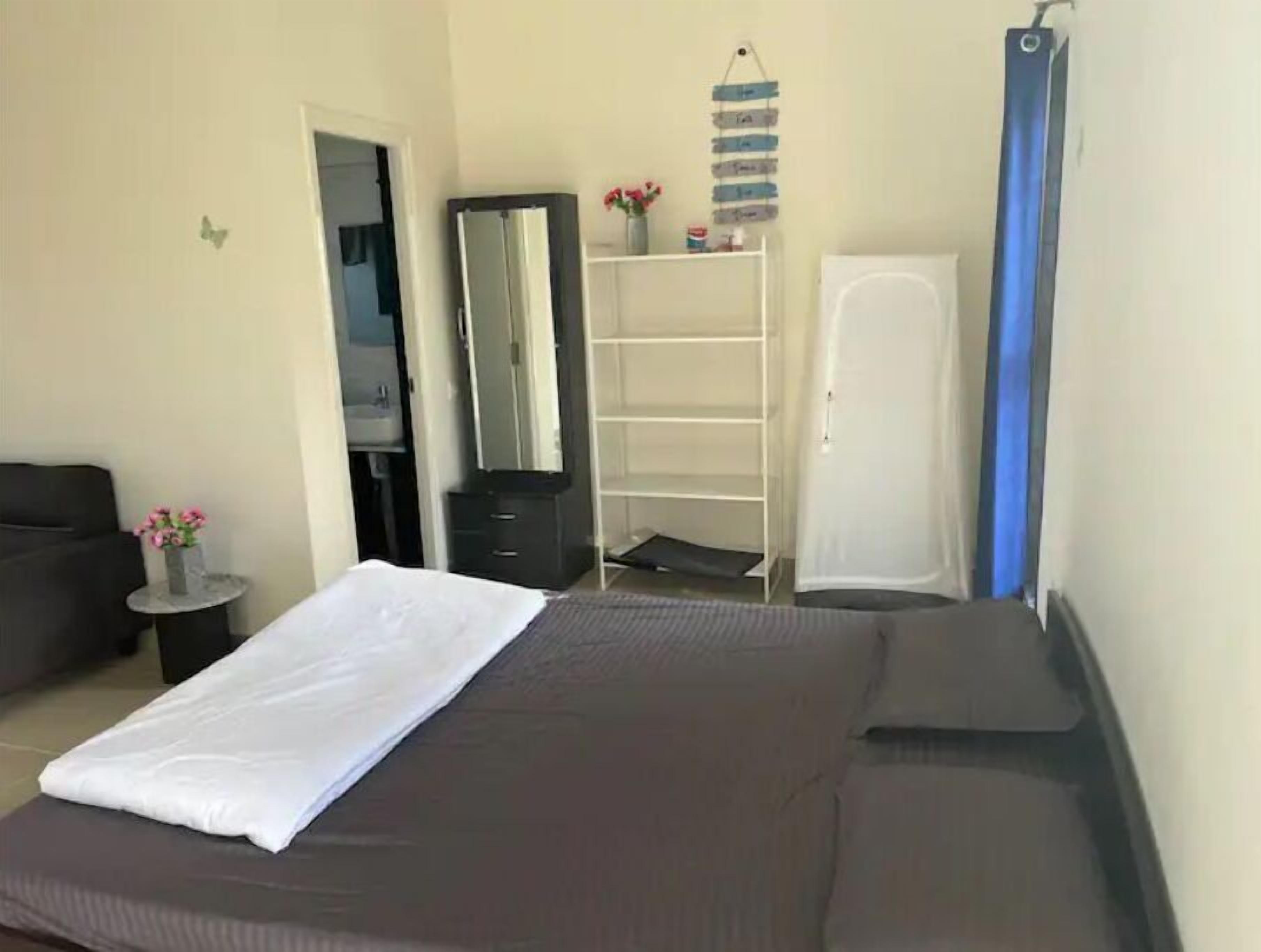 Un dormitorio con cama, armario, espejo, una mesita con un florero y una cortina azul.
