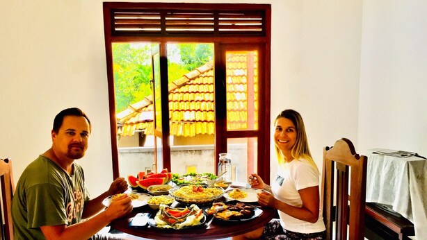 Deux personnes sont assises autour d’une table ronde, devant divers plats. On aperçoit également une chaise en bois et une table recouverte d’une nappe.