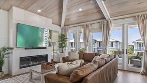 A spacious living room with a large leather sofa, a flat-screen TV mounted on the wall, a fireplace, and large windows with curtains. - 4BR New Luxury Build with Huge Private Pool + Sunset Tower Views | Esmeralda (Destin)