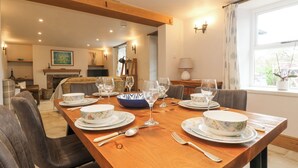 A dining area with a wooden table set for four, surrounded by chairs. A fireplace is visible in the background, and there is a window with curtains. - Woodbine Cottage (Falmouth)