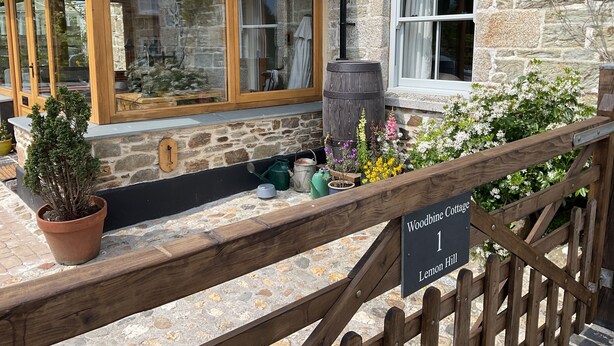 A stone cottage with wooden windows, a sign indicating "Woodbine Cottage 1 Lemon Hill," and various potted plants and gardening tools on the porch. - Woodbine Cottage (Falmouth)