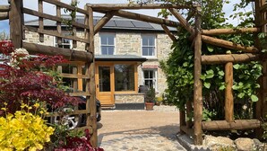 A stone building with a wooden entrance gate, a paved pathway, and various potted plants. - Woodbine Cottage (Falmouth)
