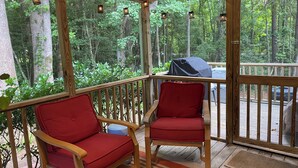 Eine überdachte Terrasse mit zwei roten Stühlen und einem Grill, mit Blick auf ein bewaldetes Gebiet.